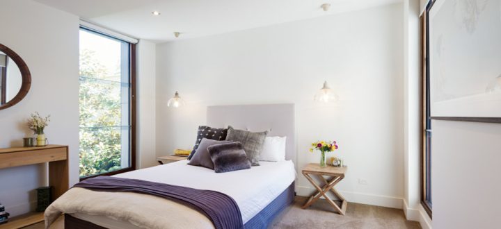 Spacious modern bedroom with large windows offering a view of green foliage outside, featuring minimalist furniture and warm lighting.