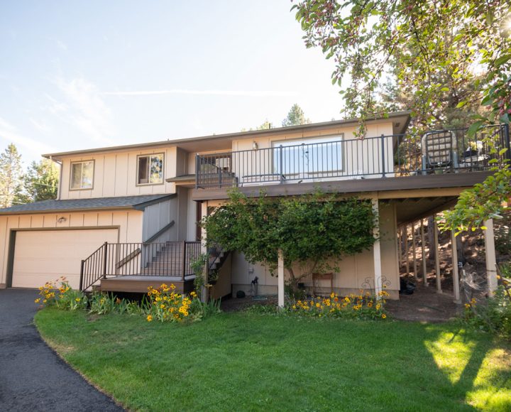 Two-tiered home exterior showing upper and lower balconies with metal railings, surrounded by green landscaping and flower beds.
