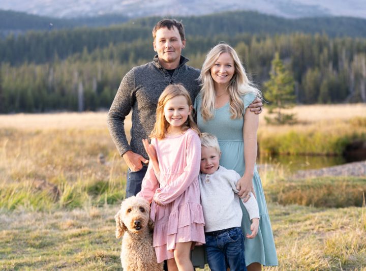 A family of four with their dog posing in a field with a mountain in the background.