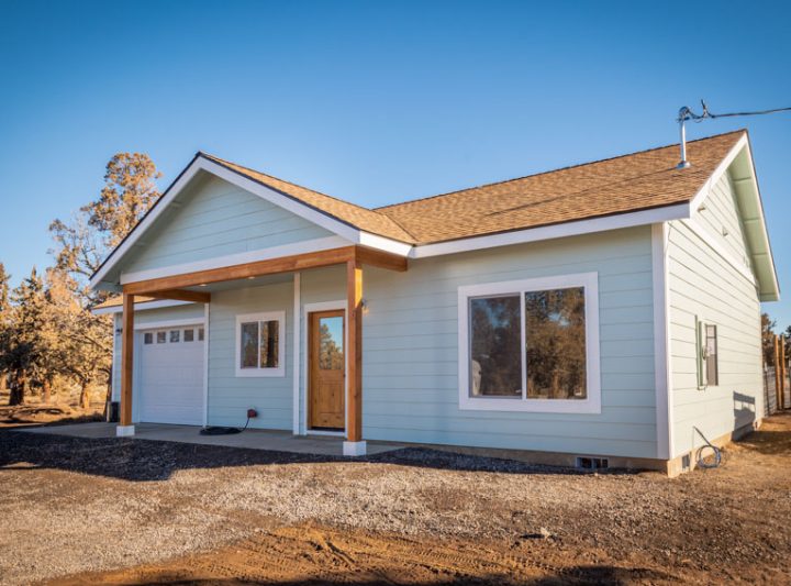Angled side-front view of a modern ADU with light blue siding, white trim, wooden porch posts, and natural surroundings—highlighting its integration into the landscape.