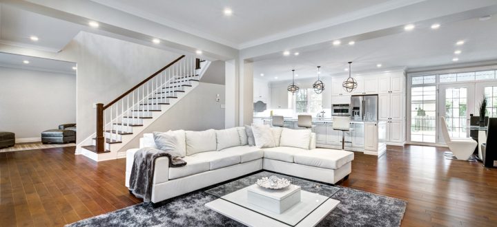 An expansive living room with a luxurious white sectional sofa, modern lighting, and an open layout kitchen with white cabinetry and dark wood flooring.